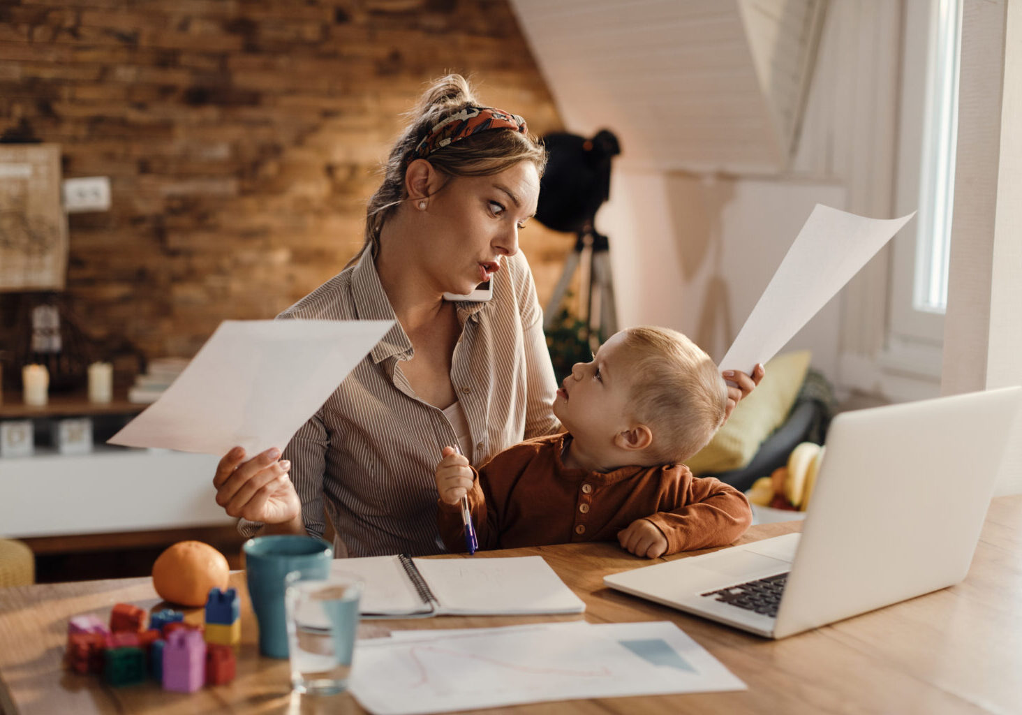 Small boy sitting on mother's lap while she is talking on cell phone and working at home.